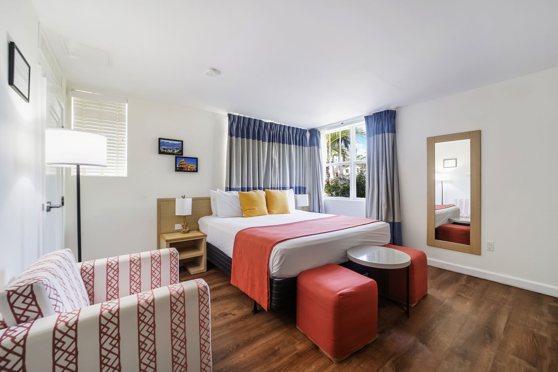 Bright hotel room with a bed, red bench, patterned chair, wall mirror, and window with striped curtains.