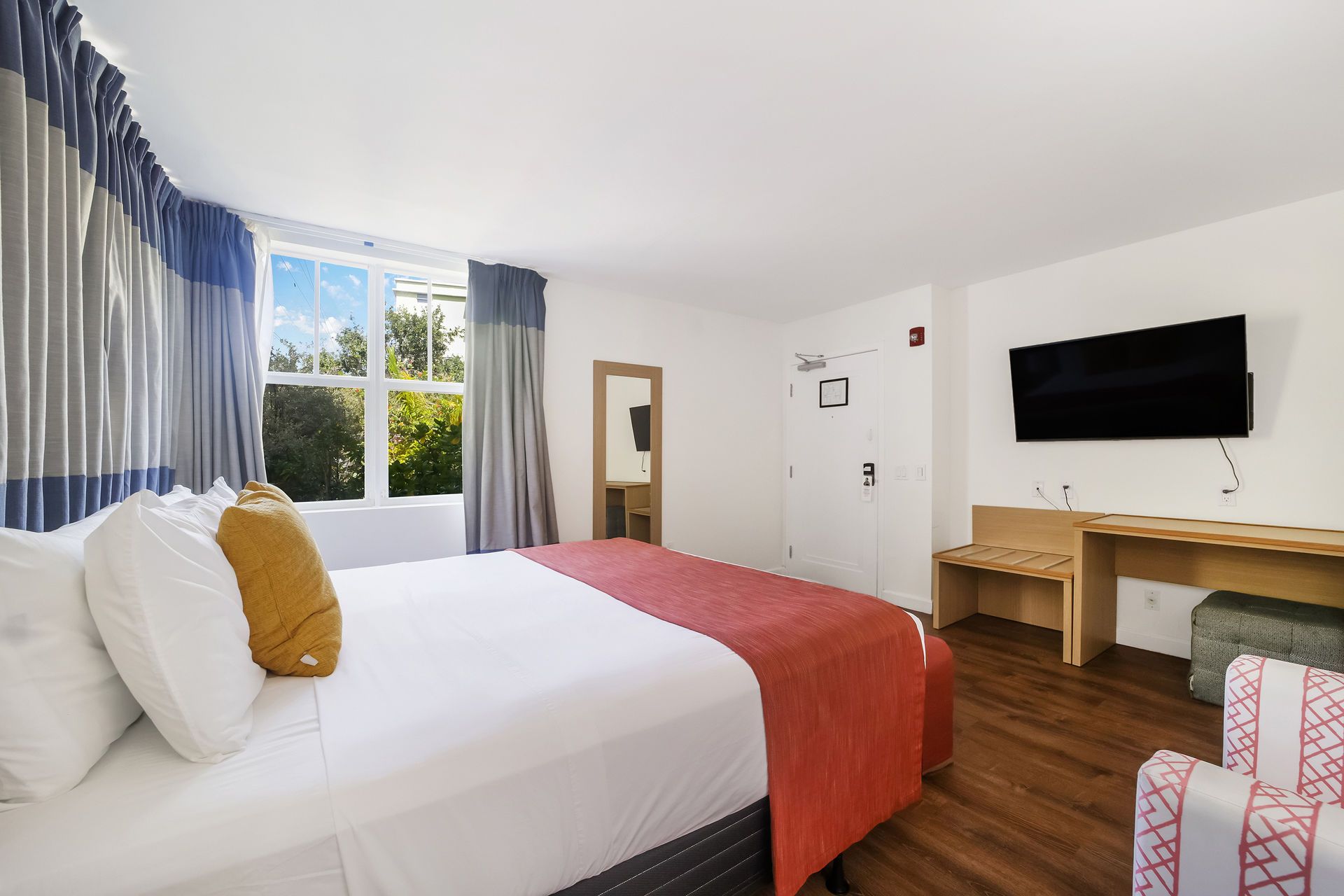 Bright hotel room with white bed, red runner, wall TV, desk, window, and patterned armchair