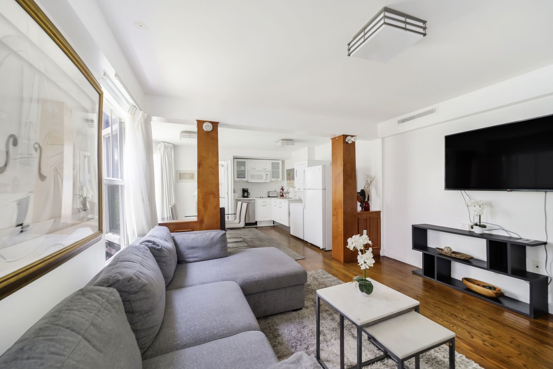Bright living room with gray sectional, hardwood floors, wall-mounted TV, and kitchen in background