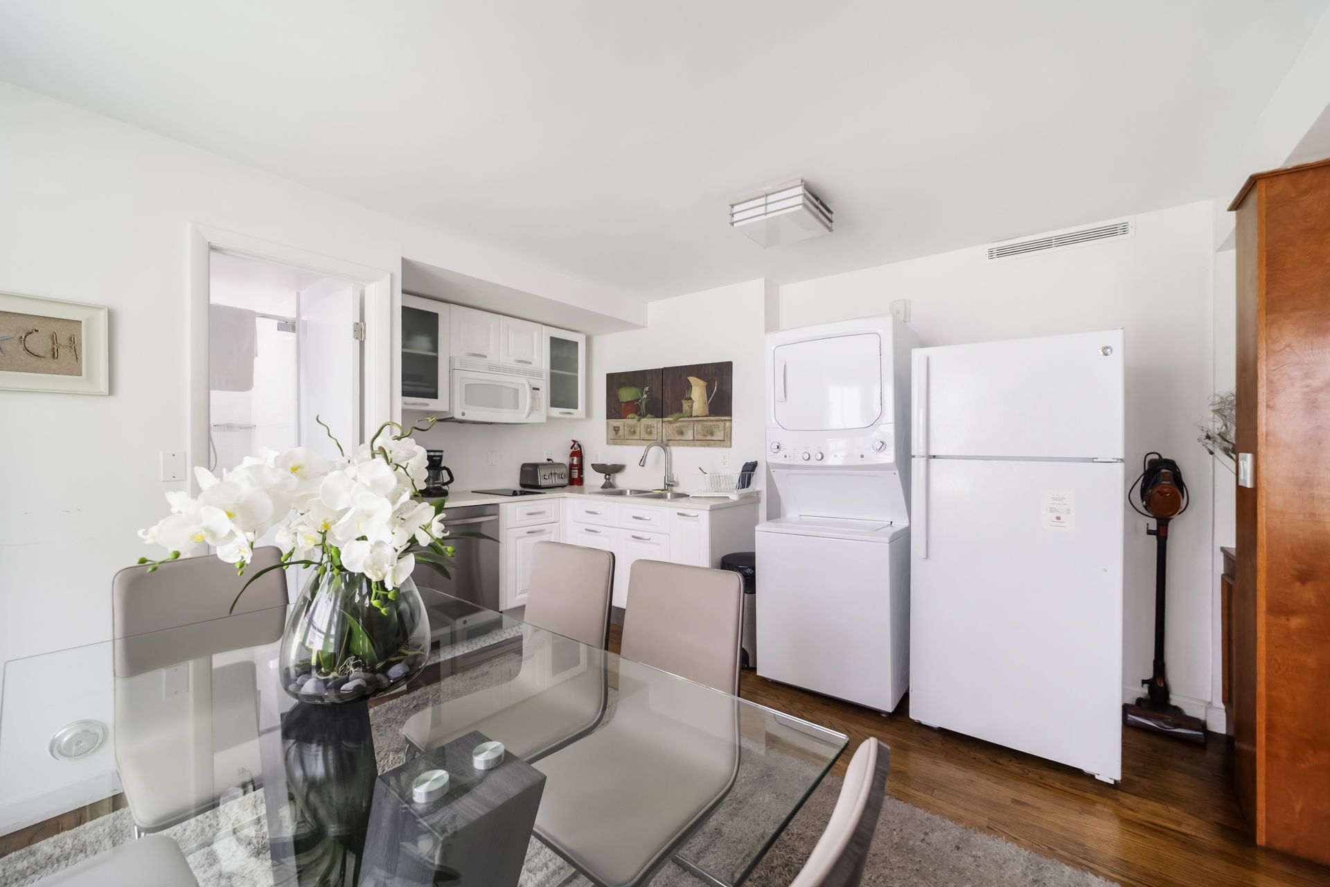 Bright dining area with glass table, white flowers, and compact kitchen with refrigerator and cabinets