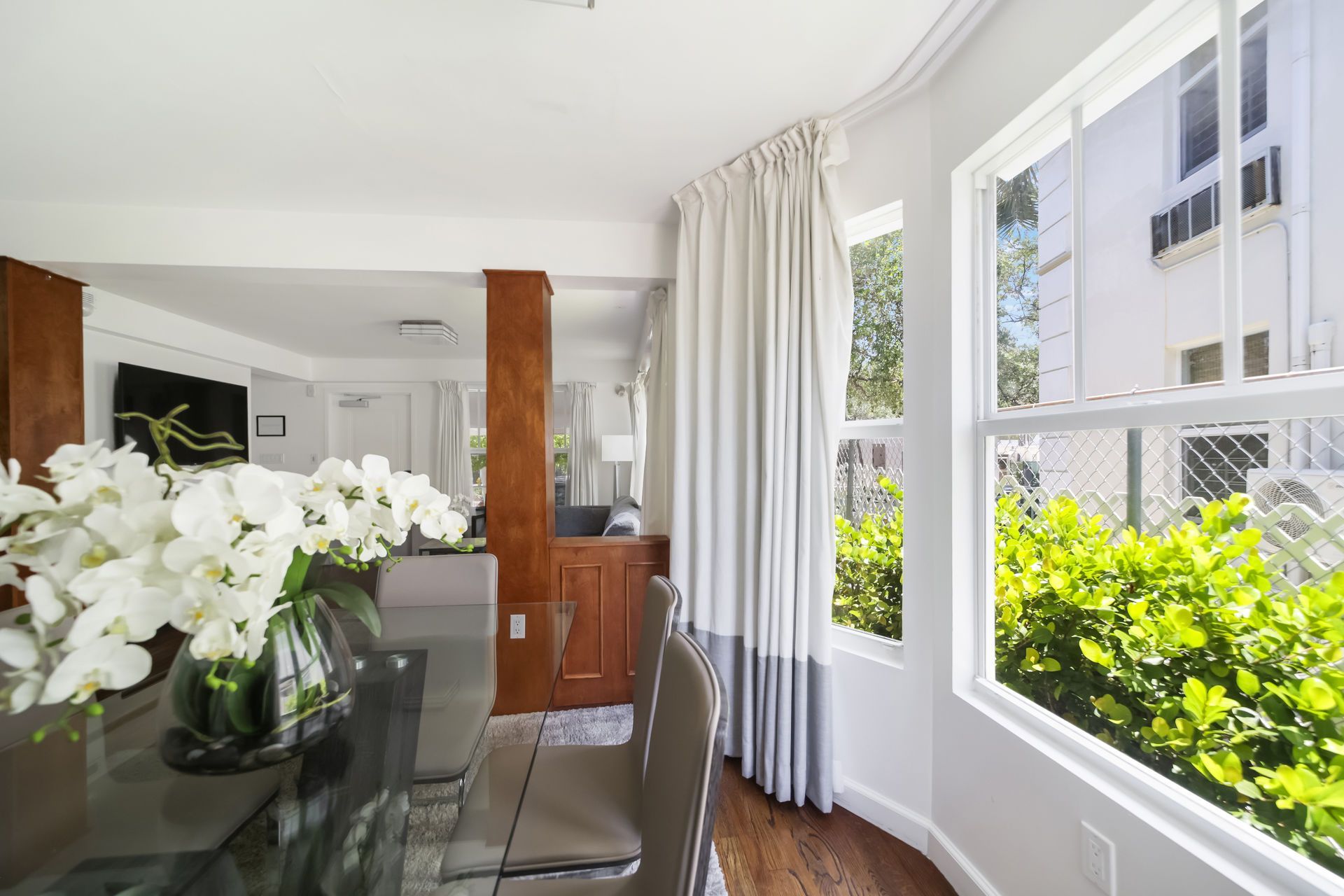 Bright dining room with glass table, white flowers, and large windows overlooking greenery