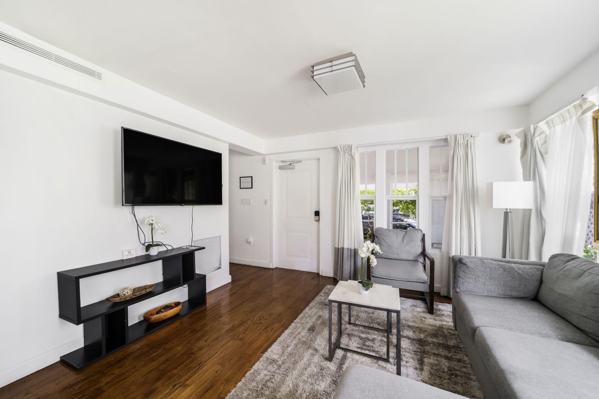Bright living room with gray sectional, wall-mounted TV, fireplace console, and large windows.