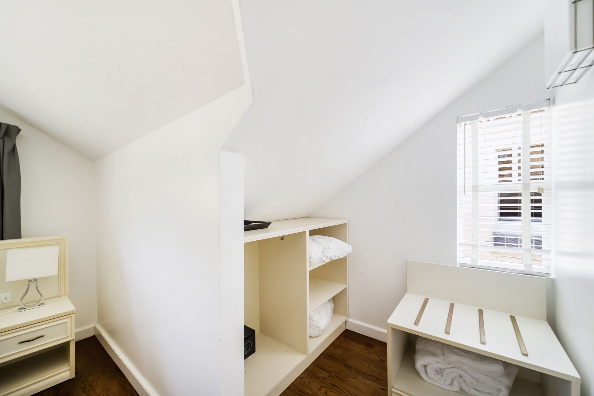 Bright white attic closet with shelves, a small vanity, and a window with blinds.