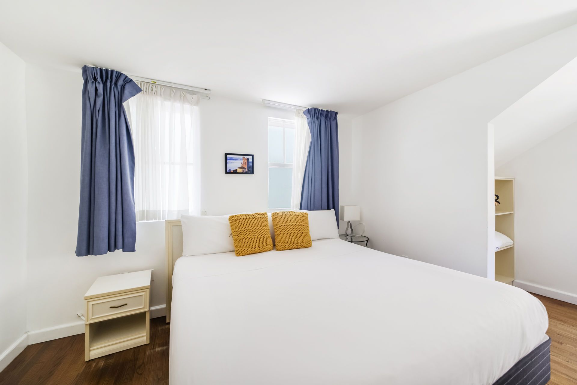 Bright bedroom with white bed, blue curtains, nightstand, and small wall art.