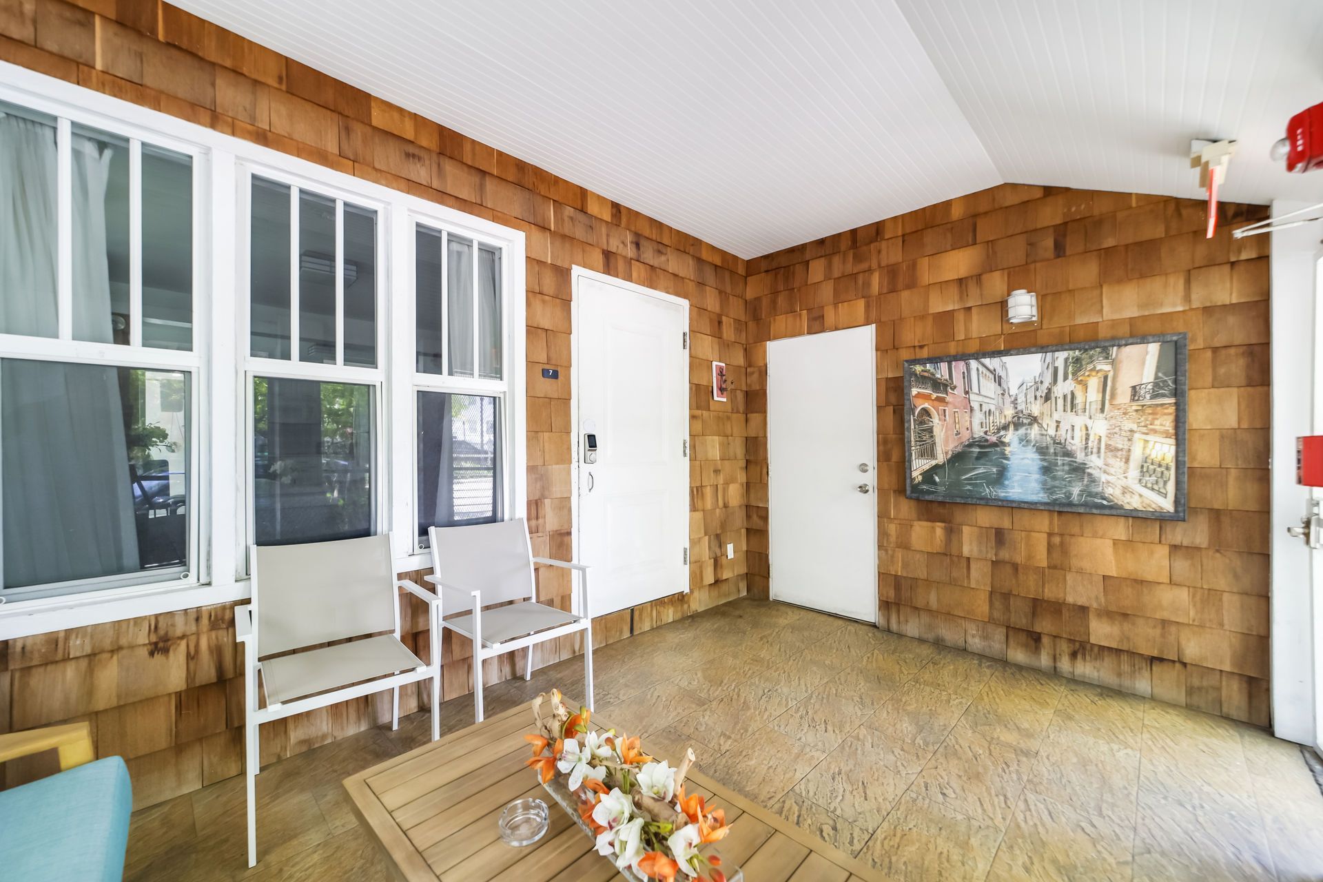 Covered porch with wood-shingle walls, white chair, large windows, and a wall painting.