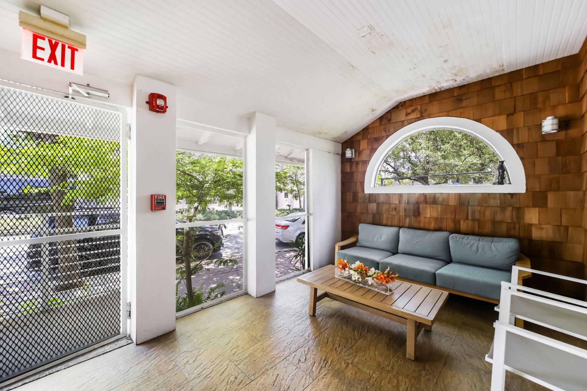 Covered patio seating area with gray sofa, coffee table, white chair, and EXIT sign beside glass door.