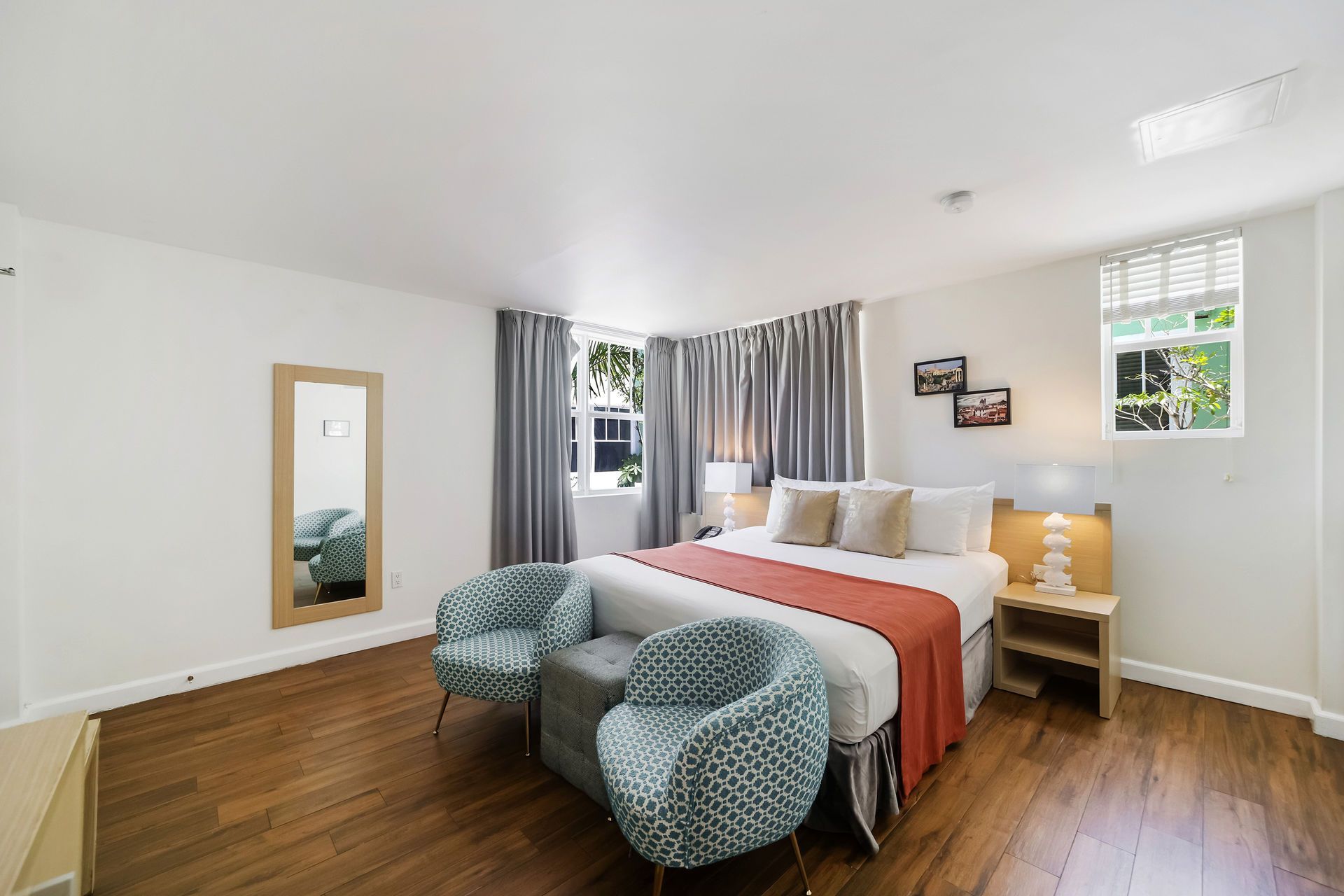Bright bedroom with white walls, wood floors, a bed with red blanket, and two teal chairs by the window.