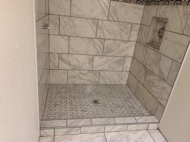 A shower stall with white tiles and a drain in a bathroom by by remodeling contractors.