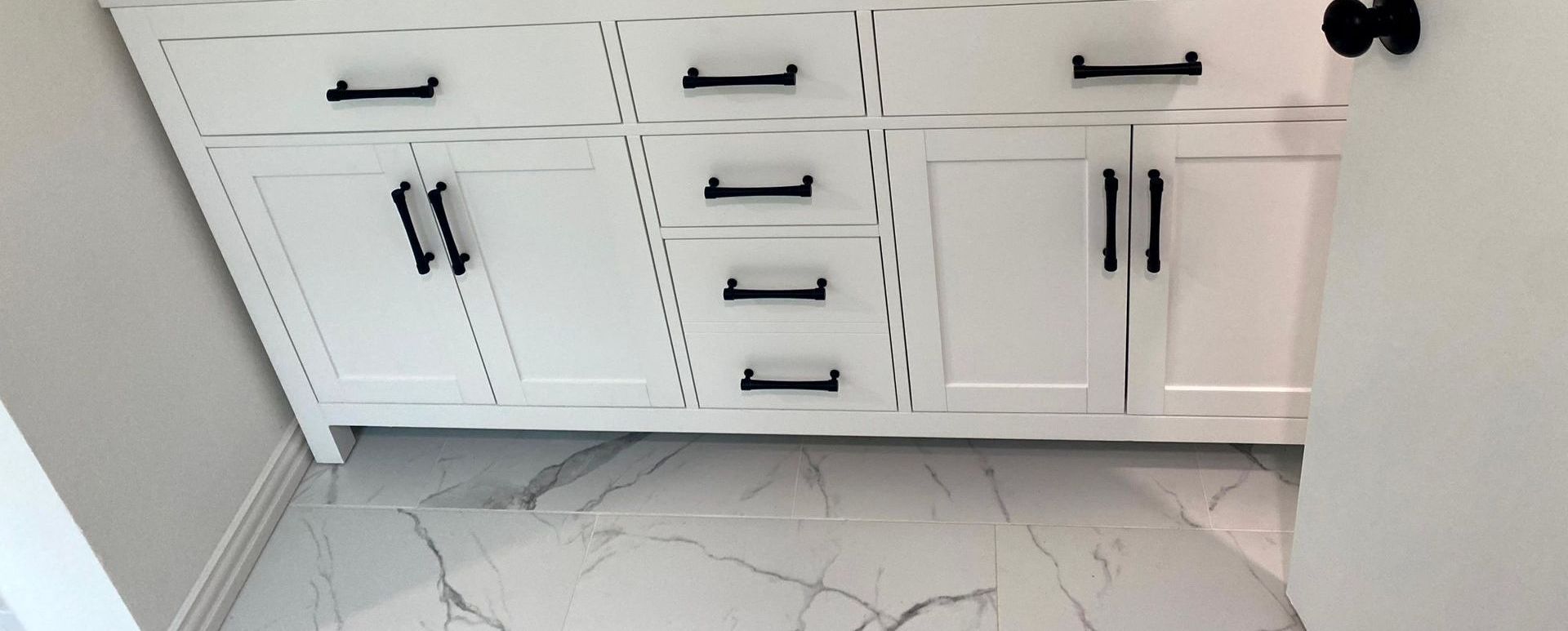 A bathroom with white cabinets and black handles and a marble floor by by remodeling contractors.