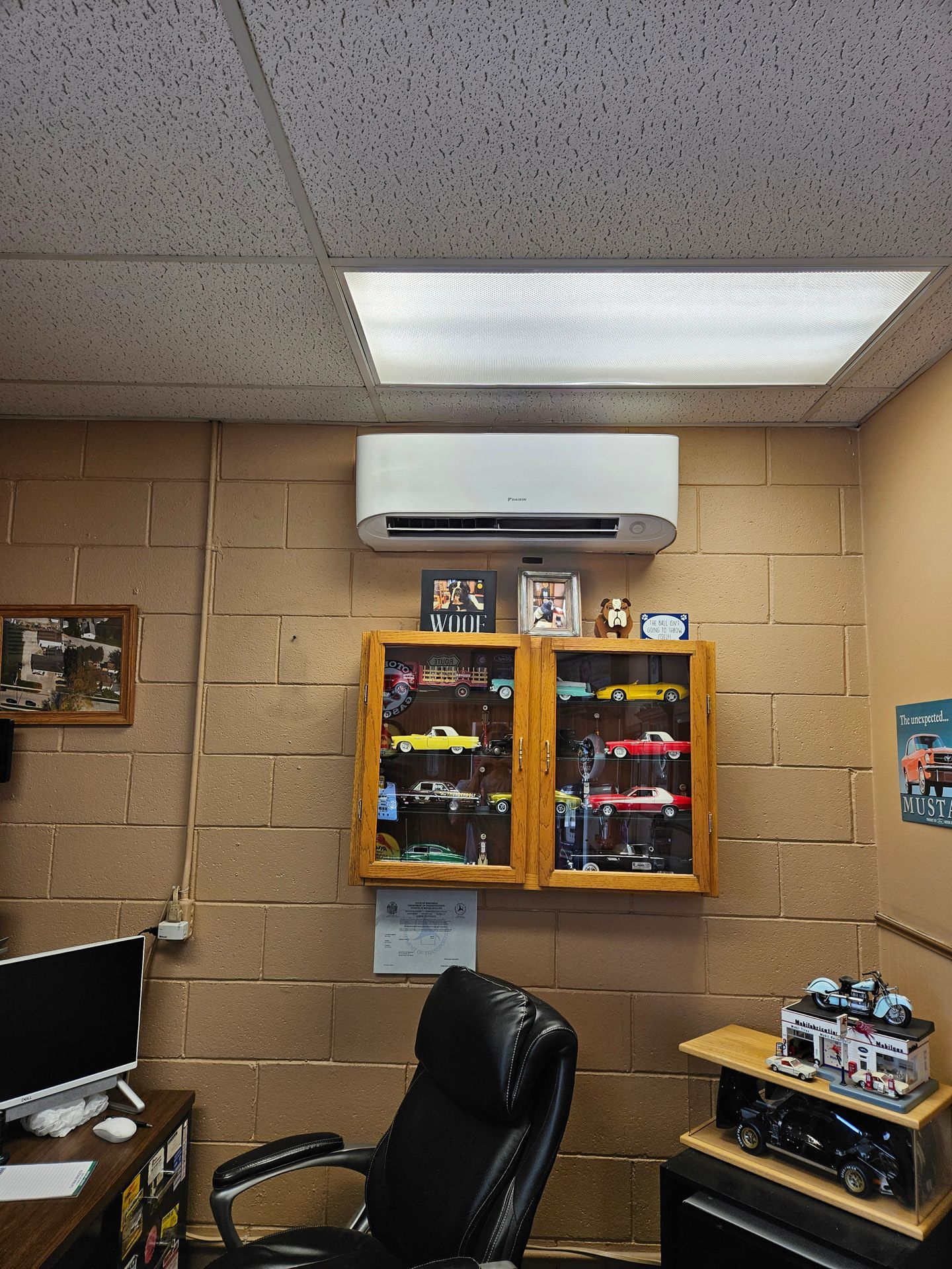 A room with air conditioner,  a chair , a computer , and a display case filled with toy cars.
