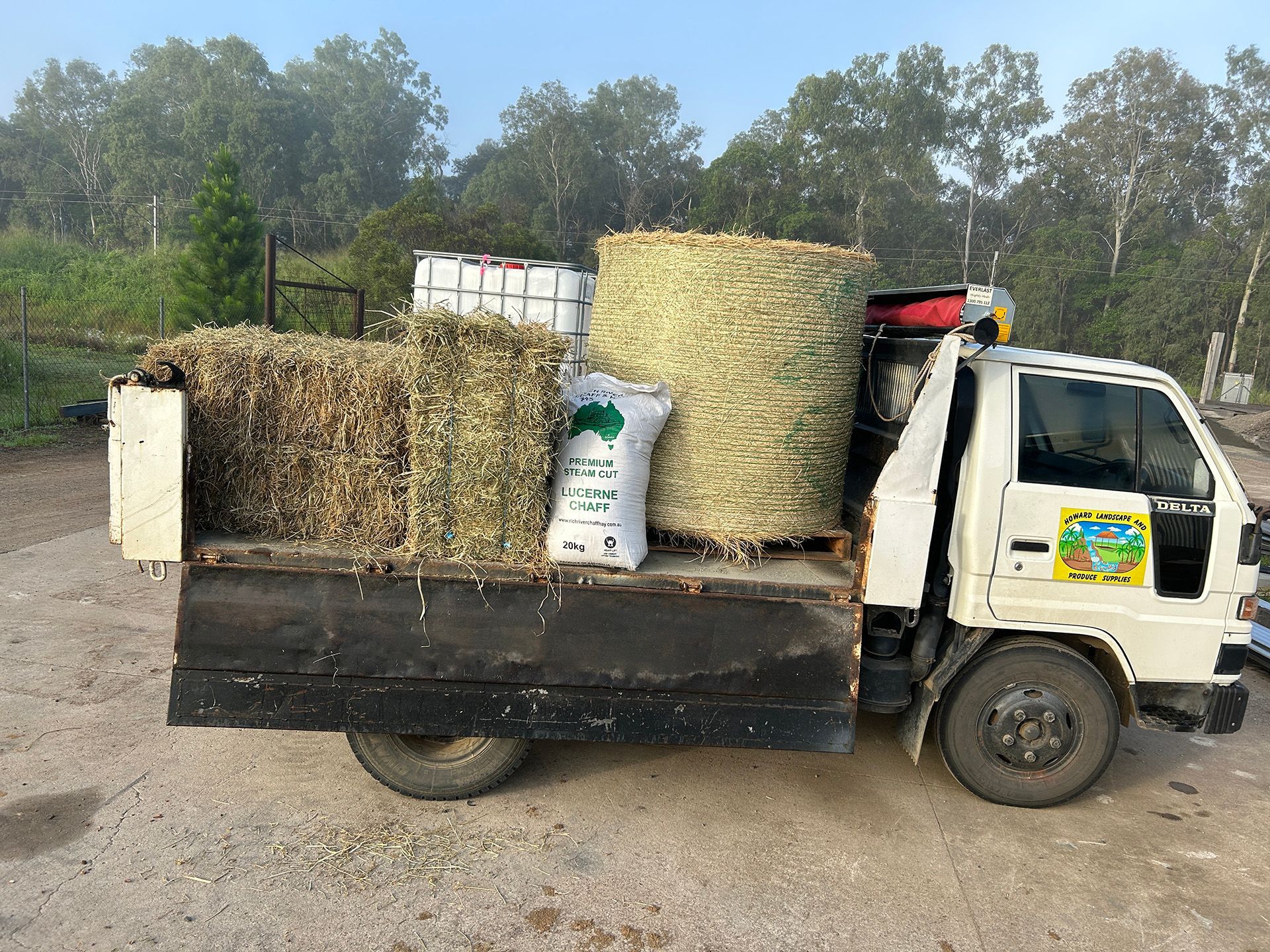 White pickup truck loaded with hay bales, feed bags, and a water tank. Outdoors, overcast day — Howard Landscape and Produce Supplies in Howard, QLD