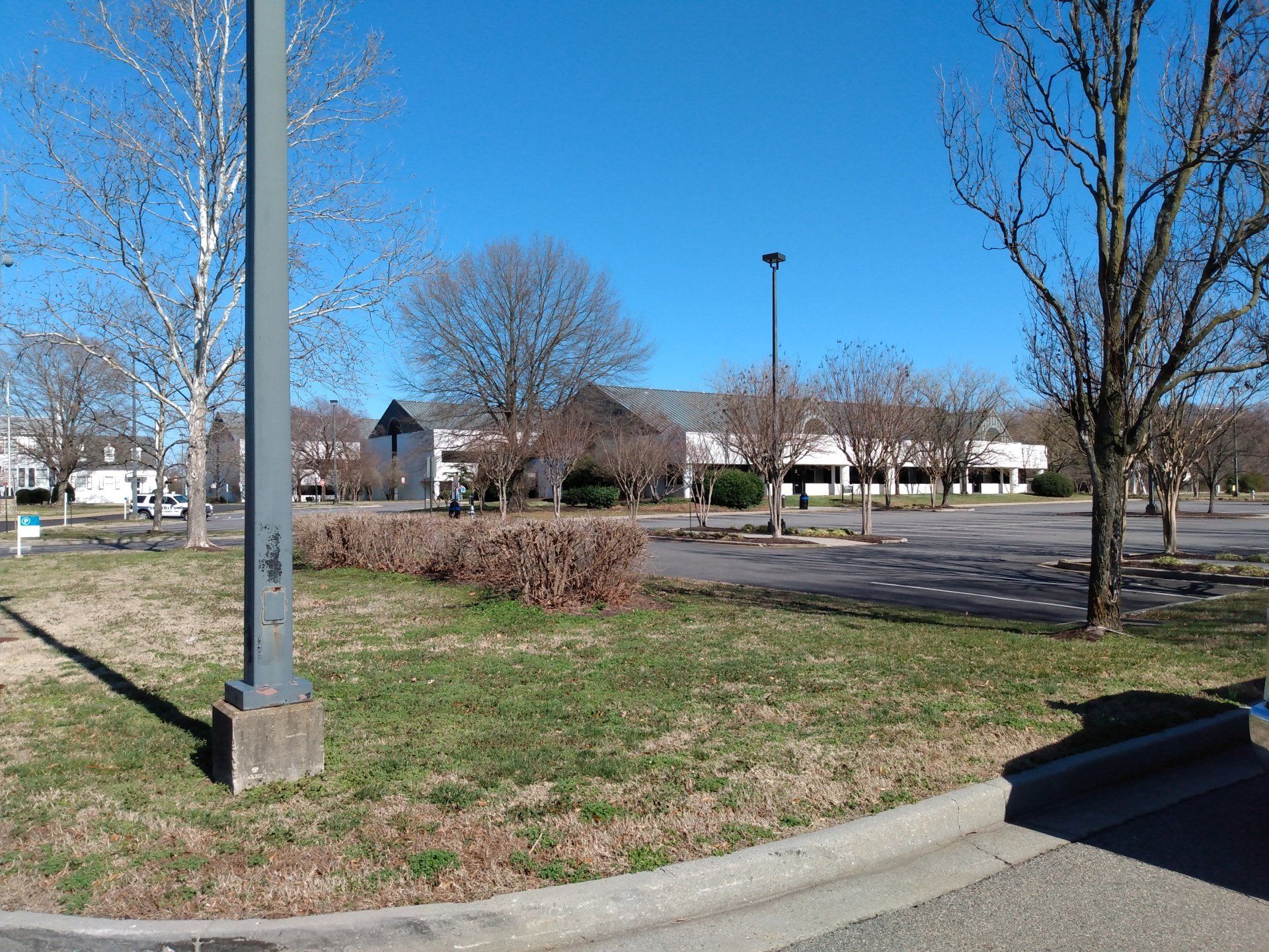 Grassy area with trees, parking lot, and a long, low building under a clear blue sky.