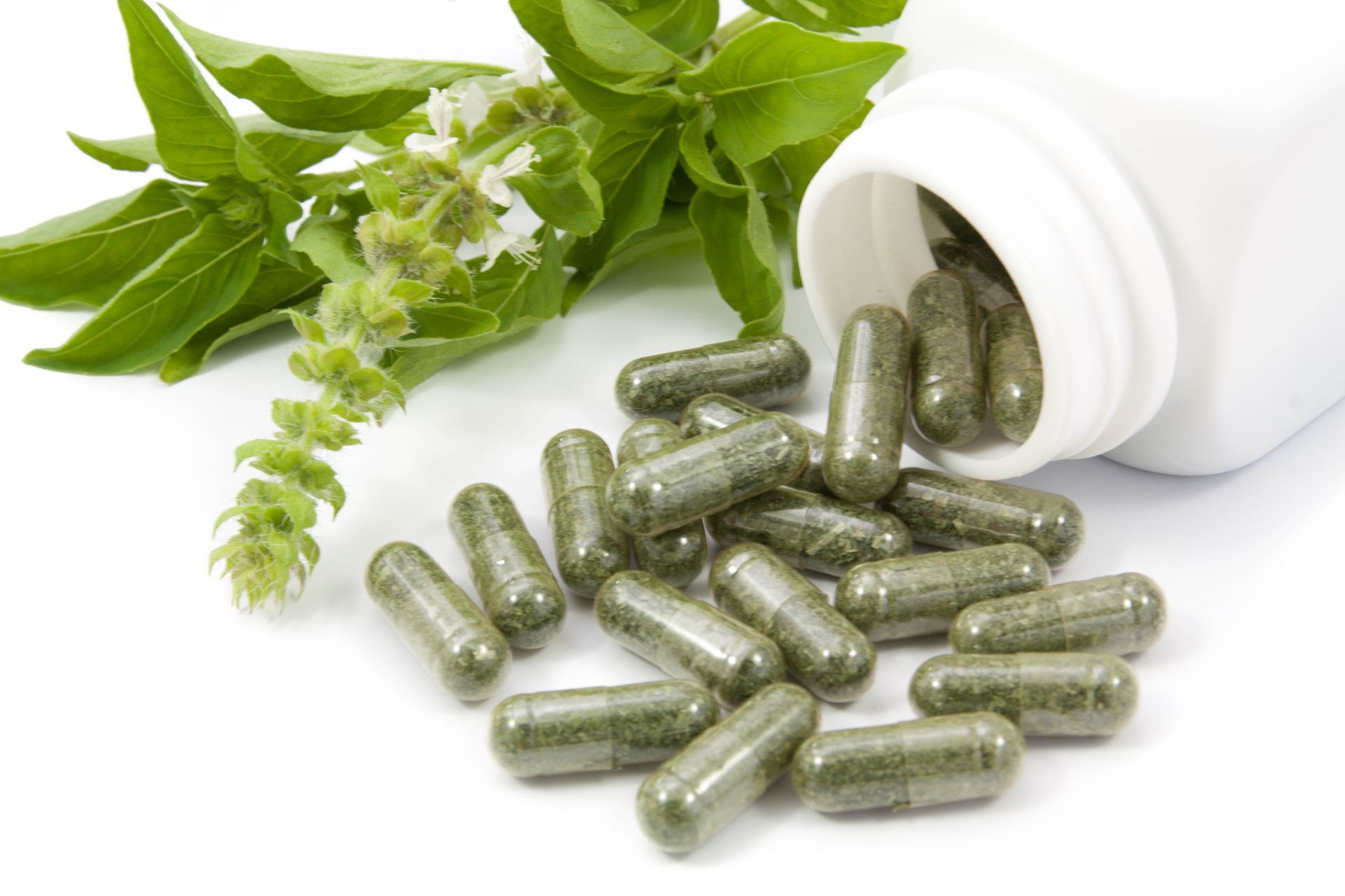 Cápsulas verdes derramándose de una botella blanca junto a una ramita de hojas y flores verdes.