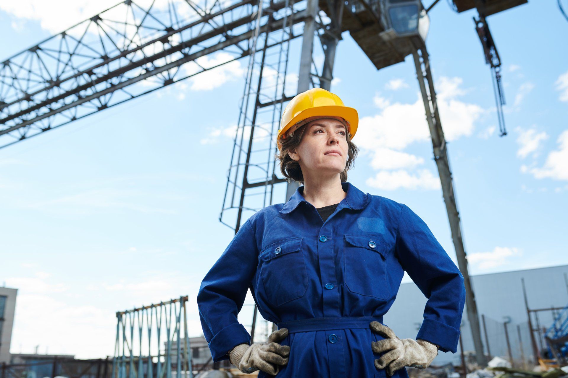 A woman wearing a hard hat and overalls is standing in front of a crane.
