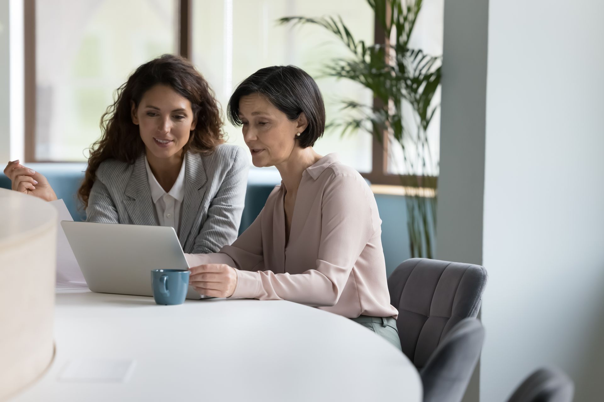 Two women looking at a laptop together, seated at a round table.