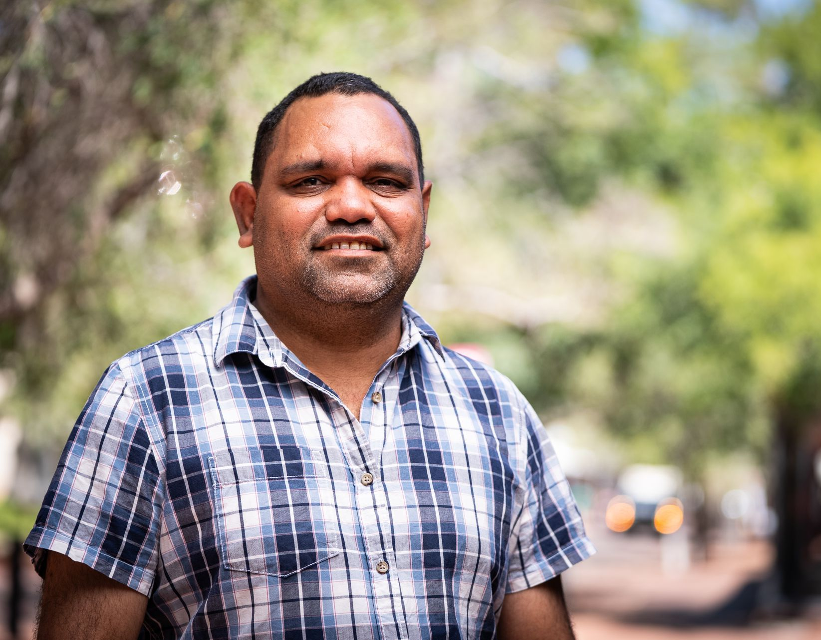 Man with brown skin wearing a blue plaid shirt smiles outdoors, trees blurred in the background.
