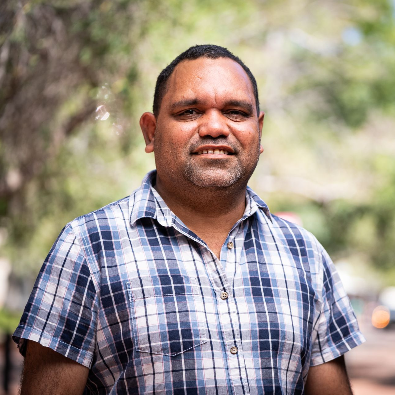 Man with dark skin wearing a blue plaid shirt smiles outdoors.
