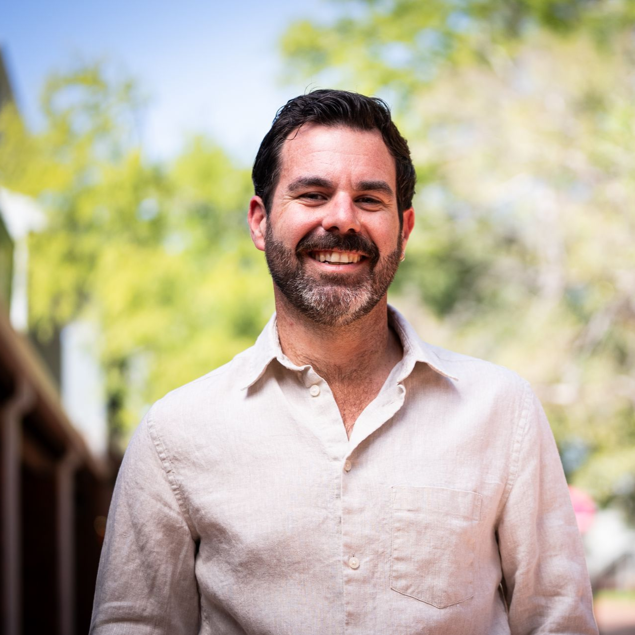 Smiling man with dark hair and beard in light shirt, outside.