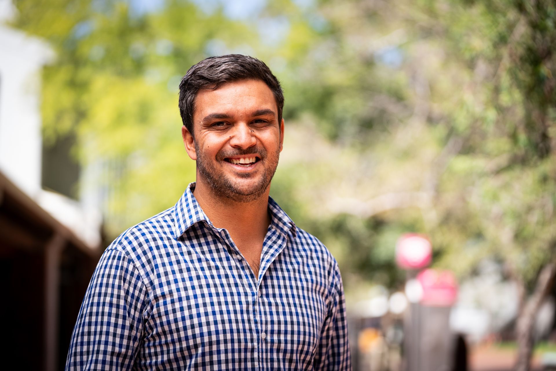 Man in a blue and white checkered shirt smiling outdoors in front of a blurred background.