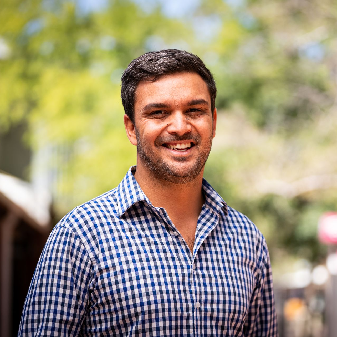 Man with dark hair and a checkered blue shirt smiles outside.