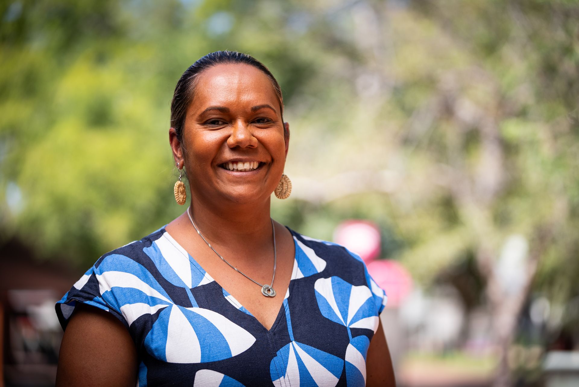 Woman smiling, wearing a blue and white patterned top, earrings, and necklace; outdoors with trees in the background.