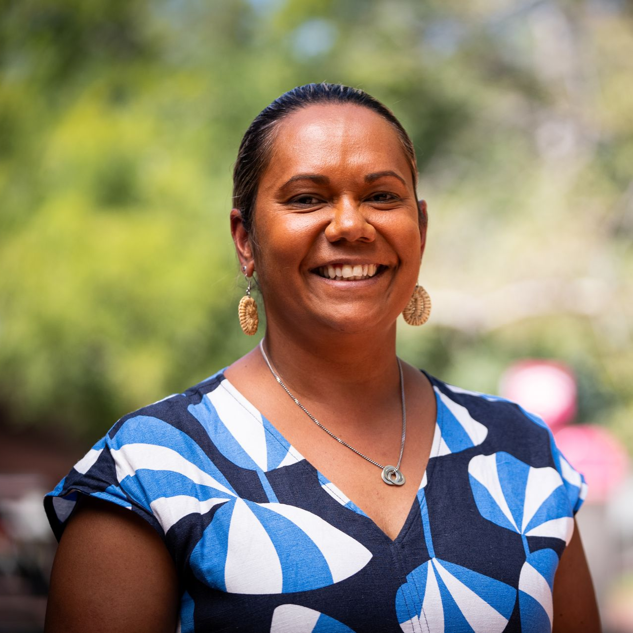 Smiling woman with dark skin, wearing a patterned blue and white top, gold earrings, and a necklace; outdoors.