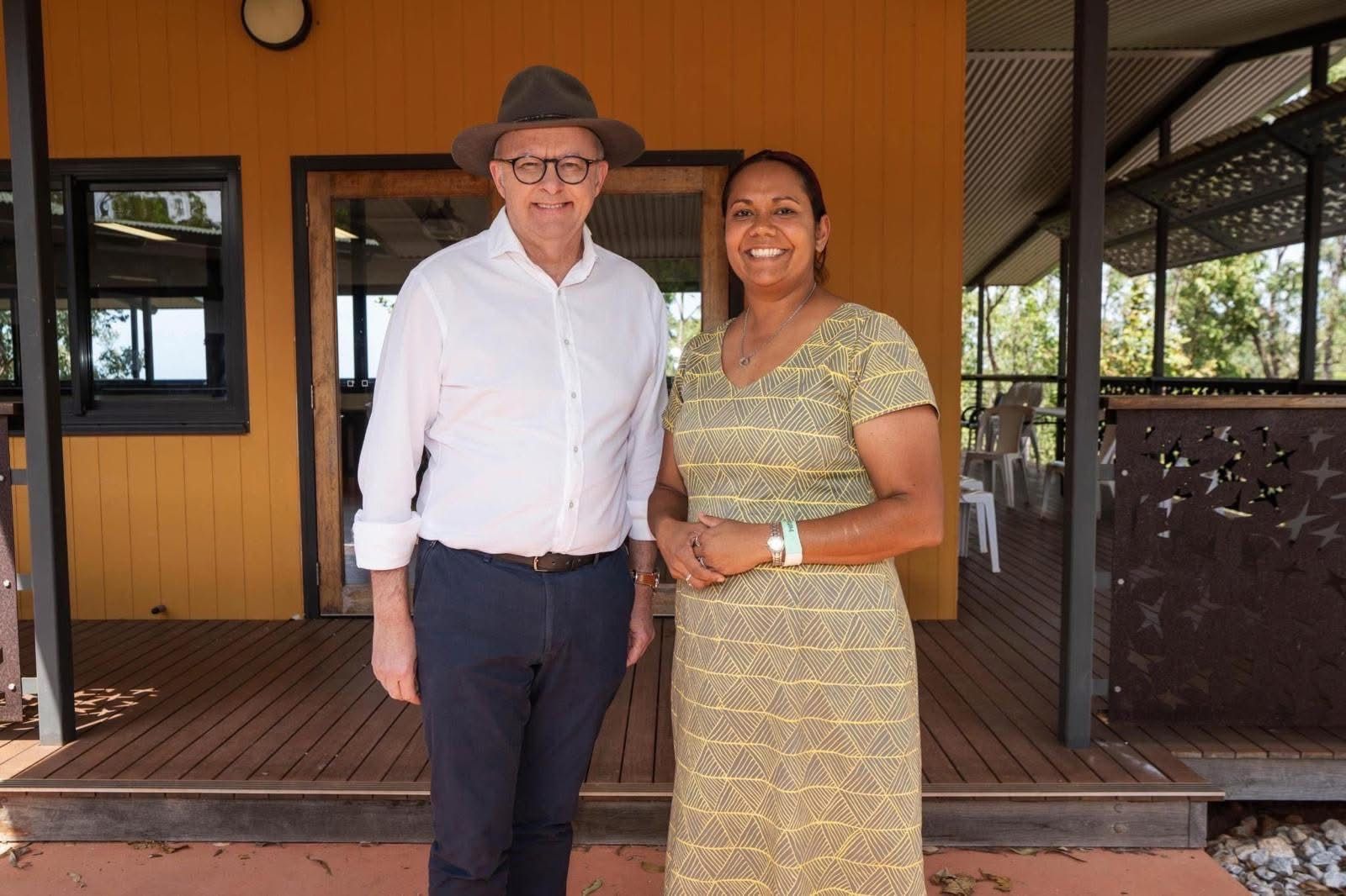Man in hat and woman smiling, standing on a wooden porch, yellow building in background.