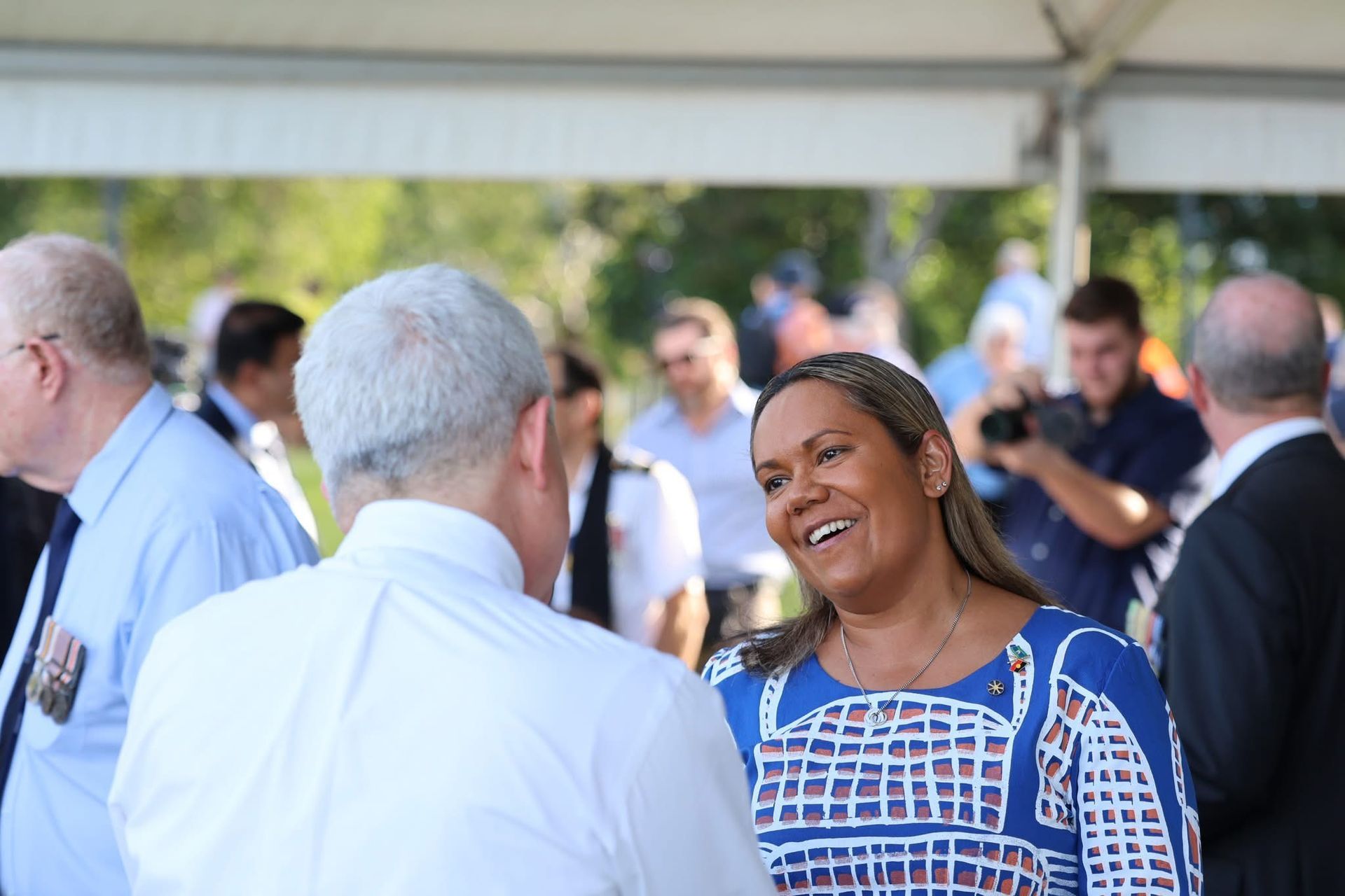 Woman smiles, talking with a man outside. Others gather nearby.