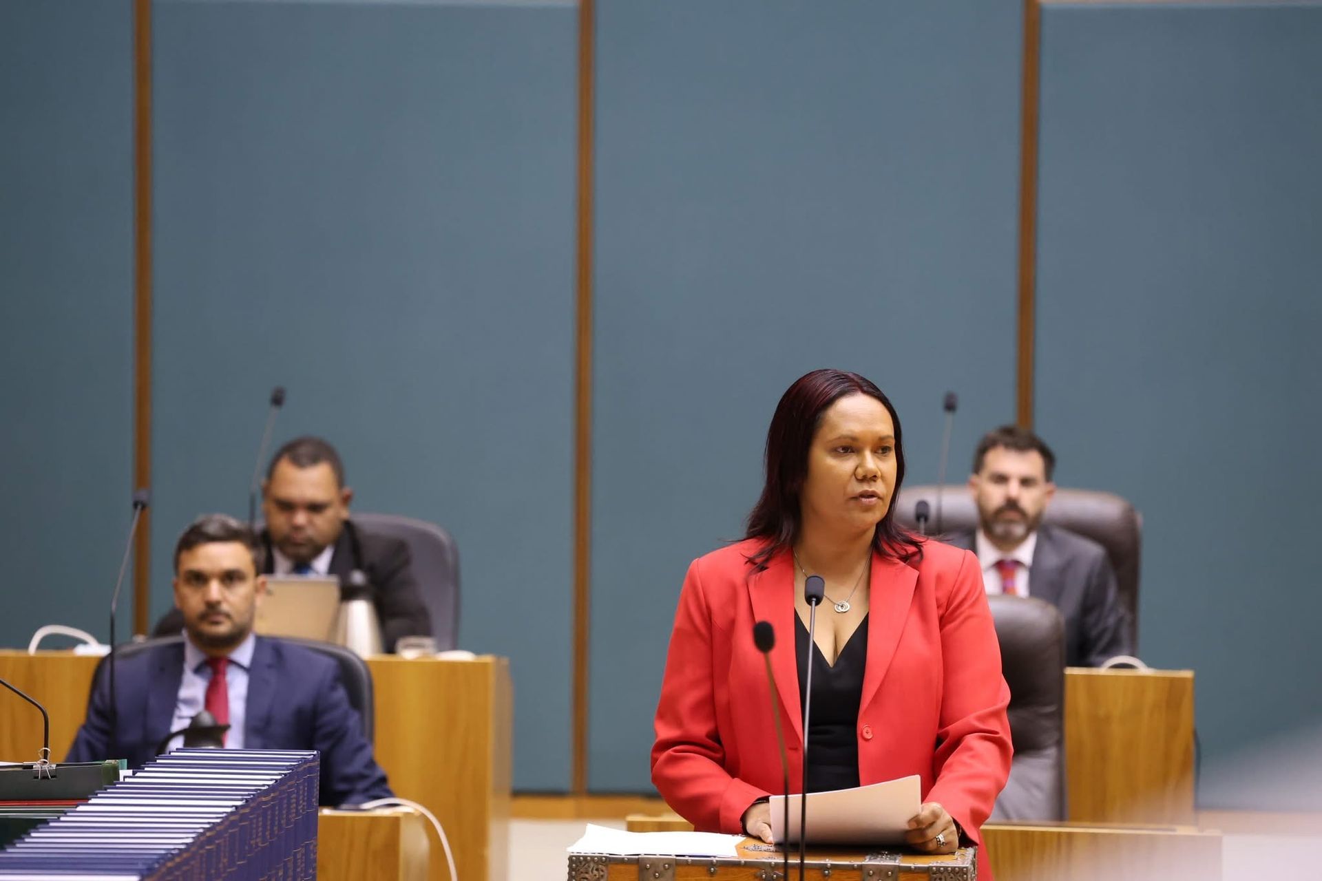 A woman in a red blazer speaks at a podium, inside a government building. Several men in suits sit in the background.