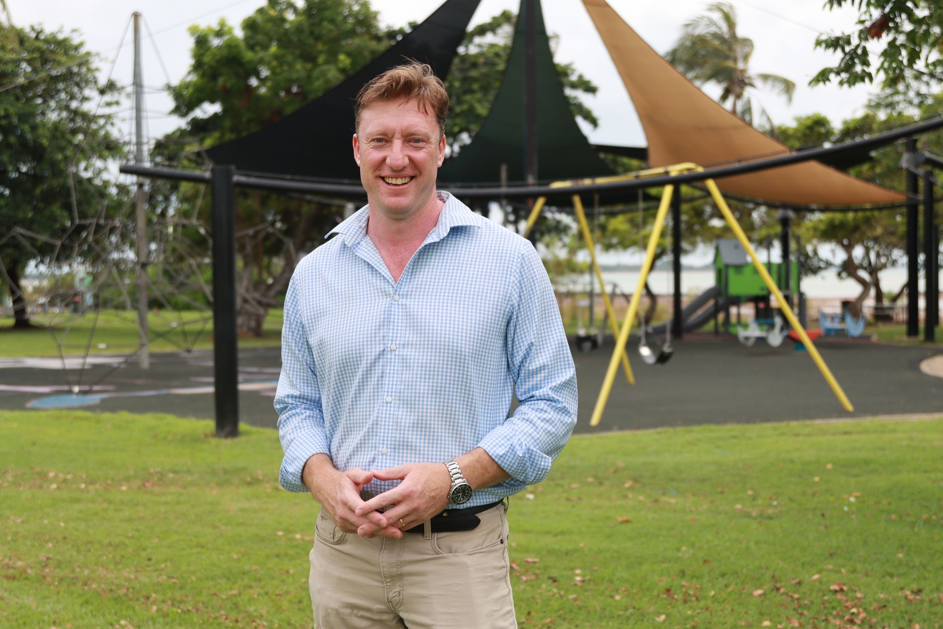 Man with a beard smiles, wearing a light-colored button-up shirt, outdoors in a green space.