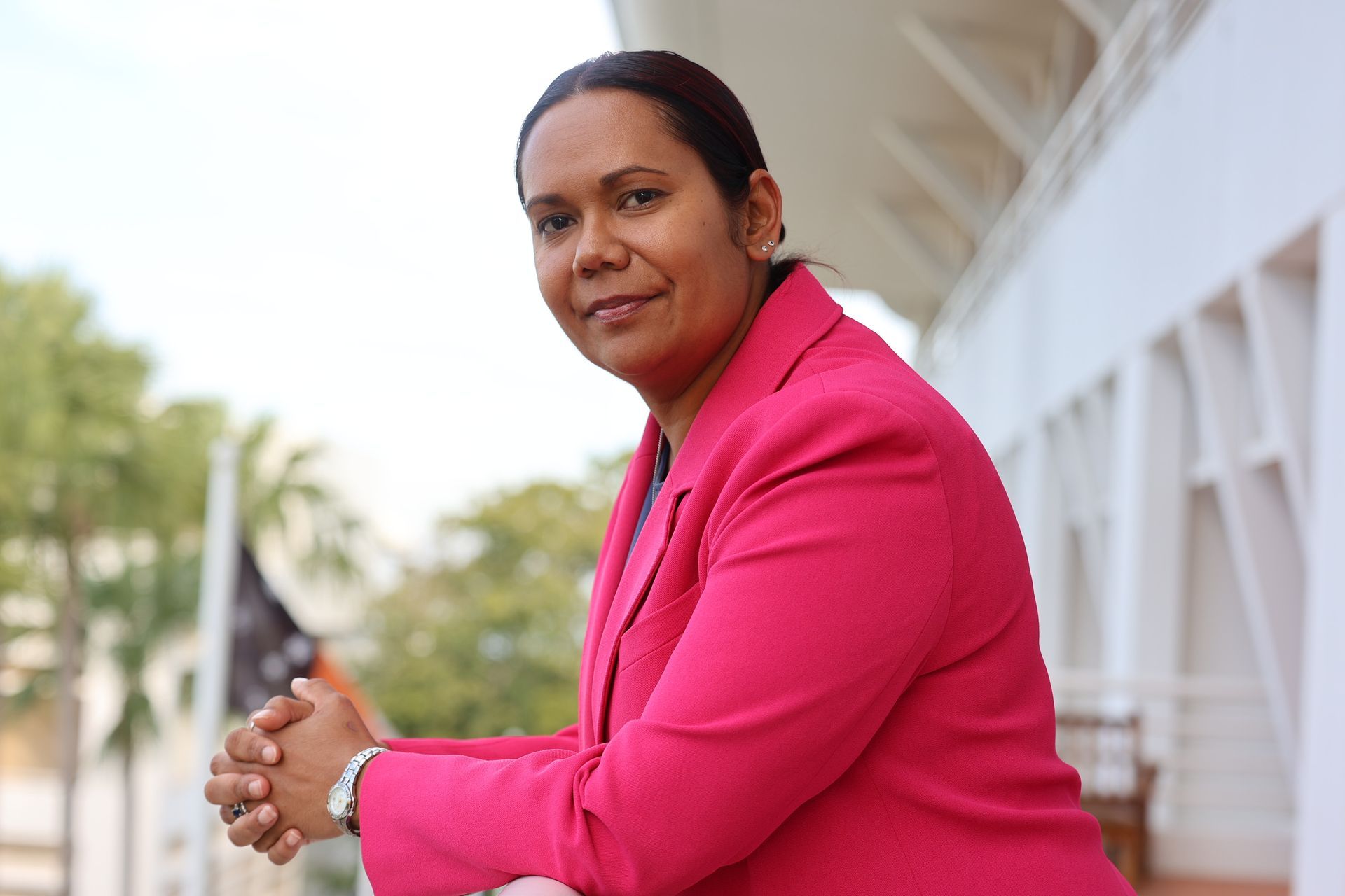 Woman in pink blazer, arms folded, leans on a balcony. Bright sunlight.
