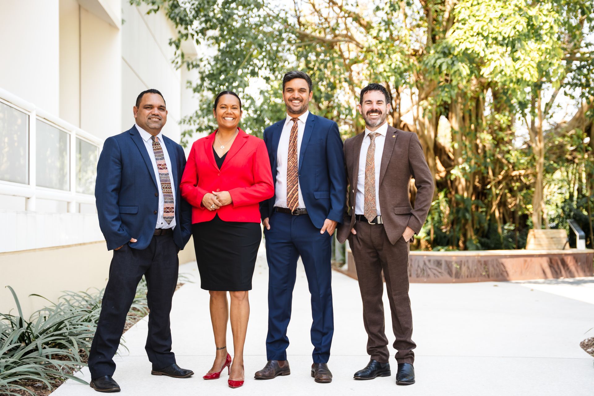 Four diverse professionals standing outdoors, smiling. One in a red jacket, others in suits with ties.