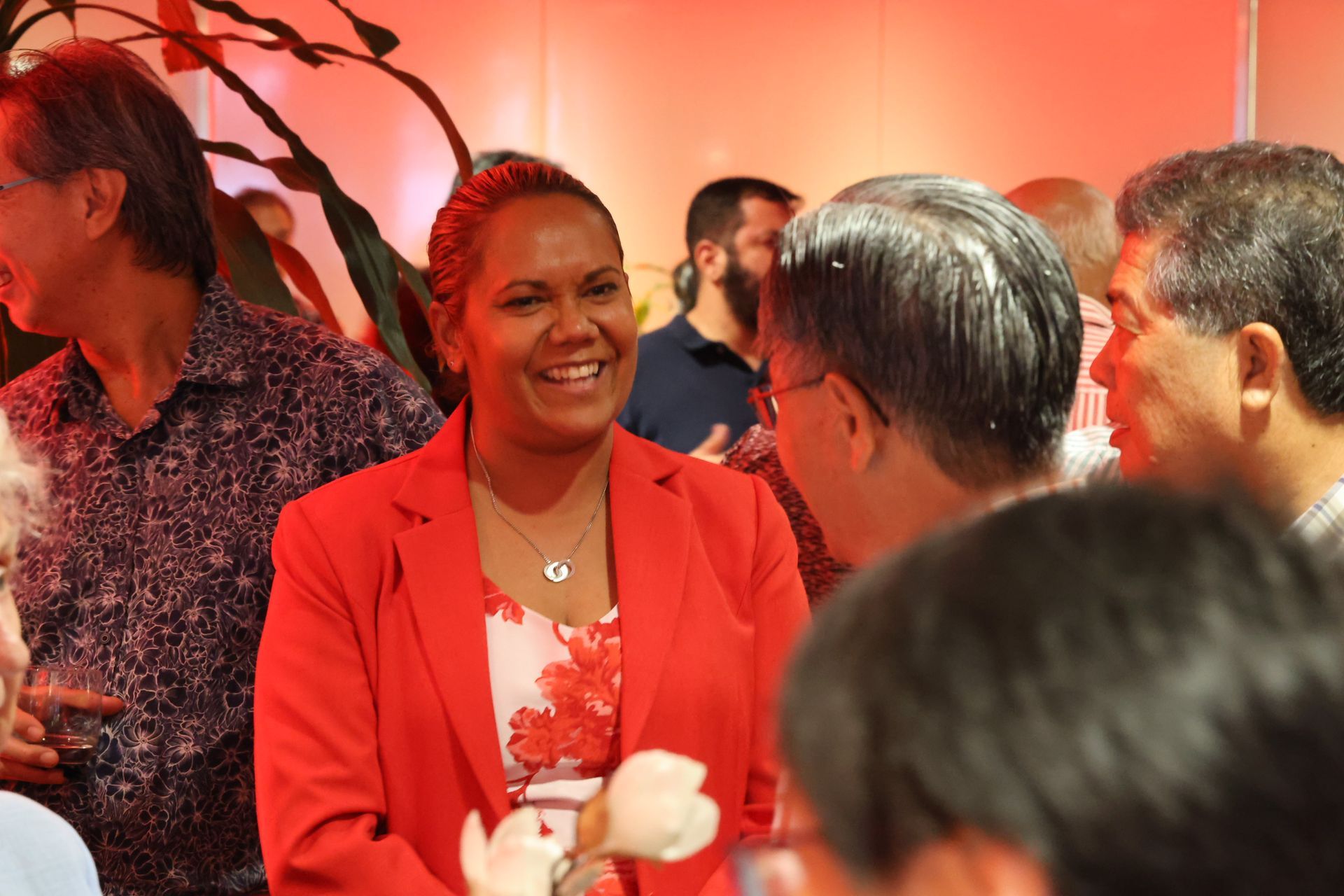 Woman in red blazer smiles, talking to a group of people in a brightly lit room.