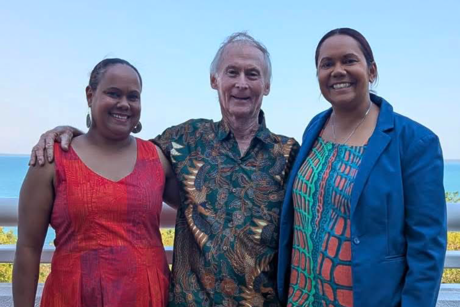 Three people smiling, posing outside with the ocean in the background.
