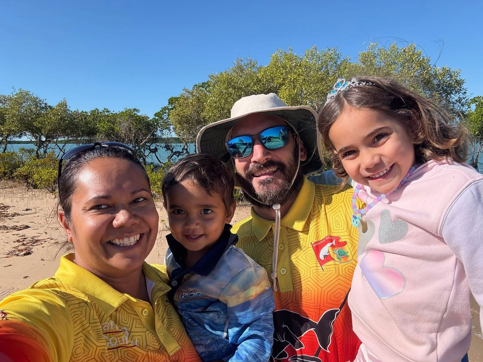 Family smiling for a selfie on a sunny beach. Includes two adults and two children with colorful shirts.