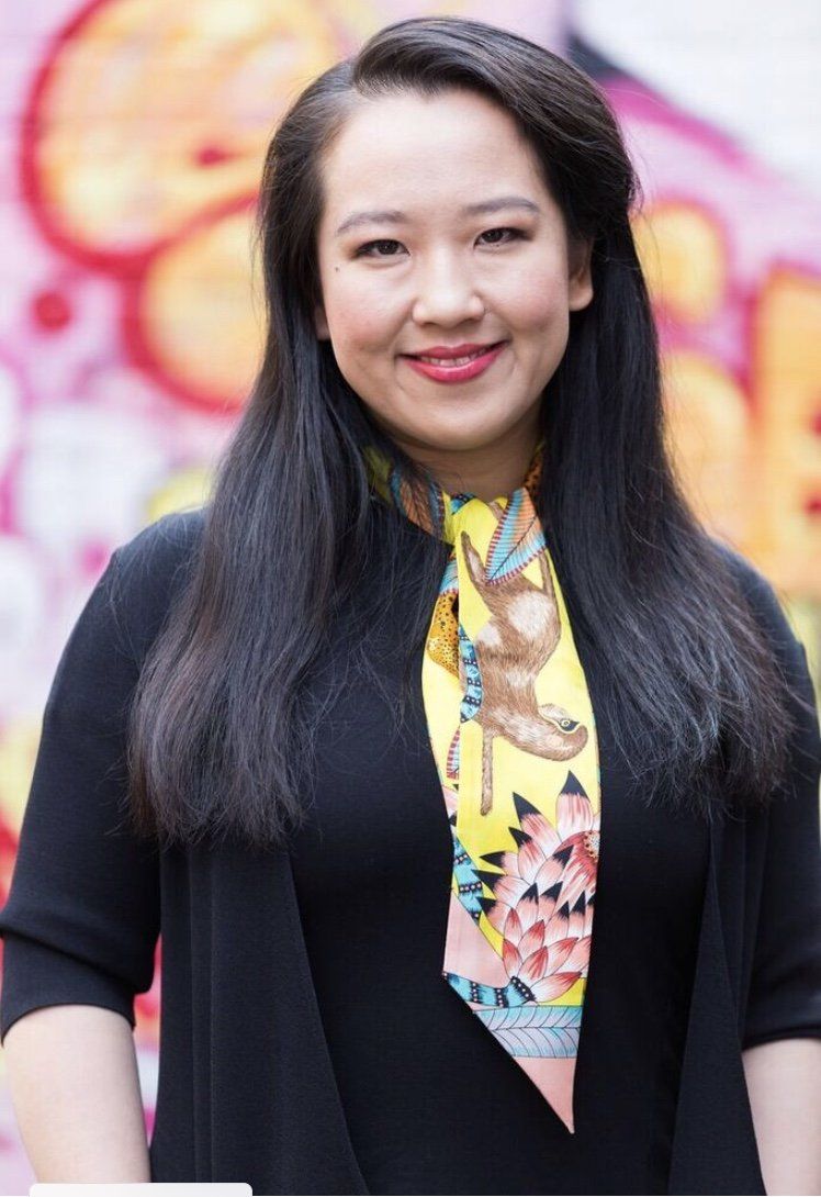 A woman wearing a black jacket and a yellow scarf is standing in front of a graffiti wall.