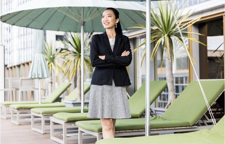 A woman is standing in front of a row of green lounge chairs under an umbrella.