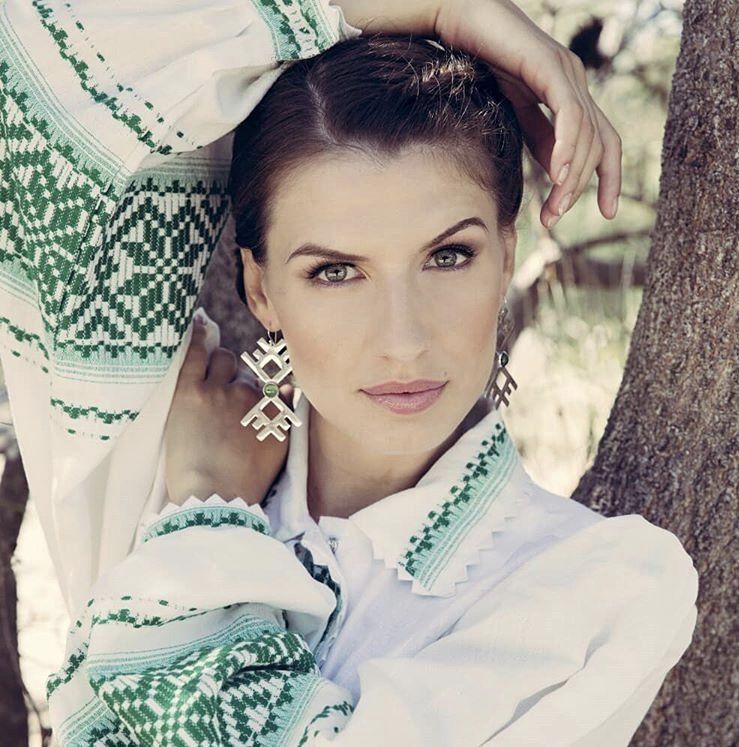A woman wearing a white shirt with green embroidery is standing next to a tree