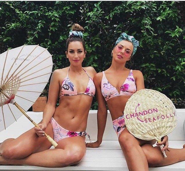 Two women in bikinis sit on a bench holding umbrellas and a fan that says chandon seafolly