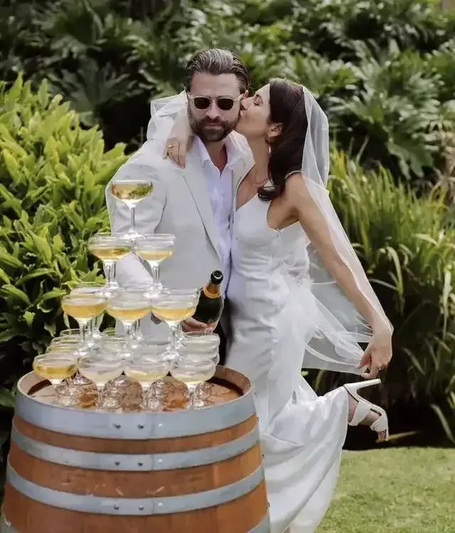 A bride and groom are kissing in front of a tower of champagne glasses.