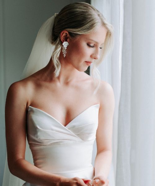 A woman in a white dress and veil is standing in front of a window.