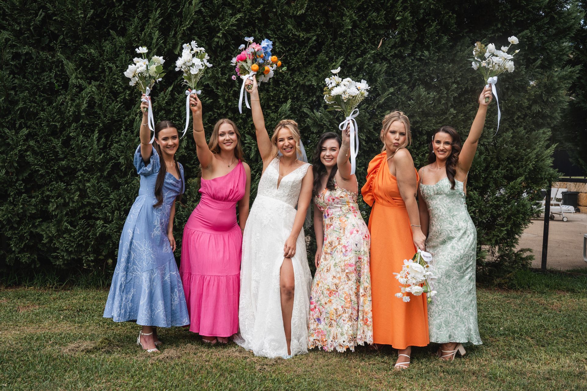 bride and her colourfully dressed bridesmaids holding bouquets of flowers in the air after having hair and makeup done