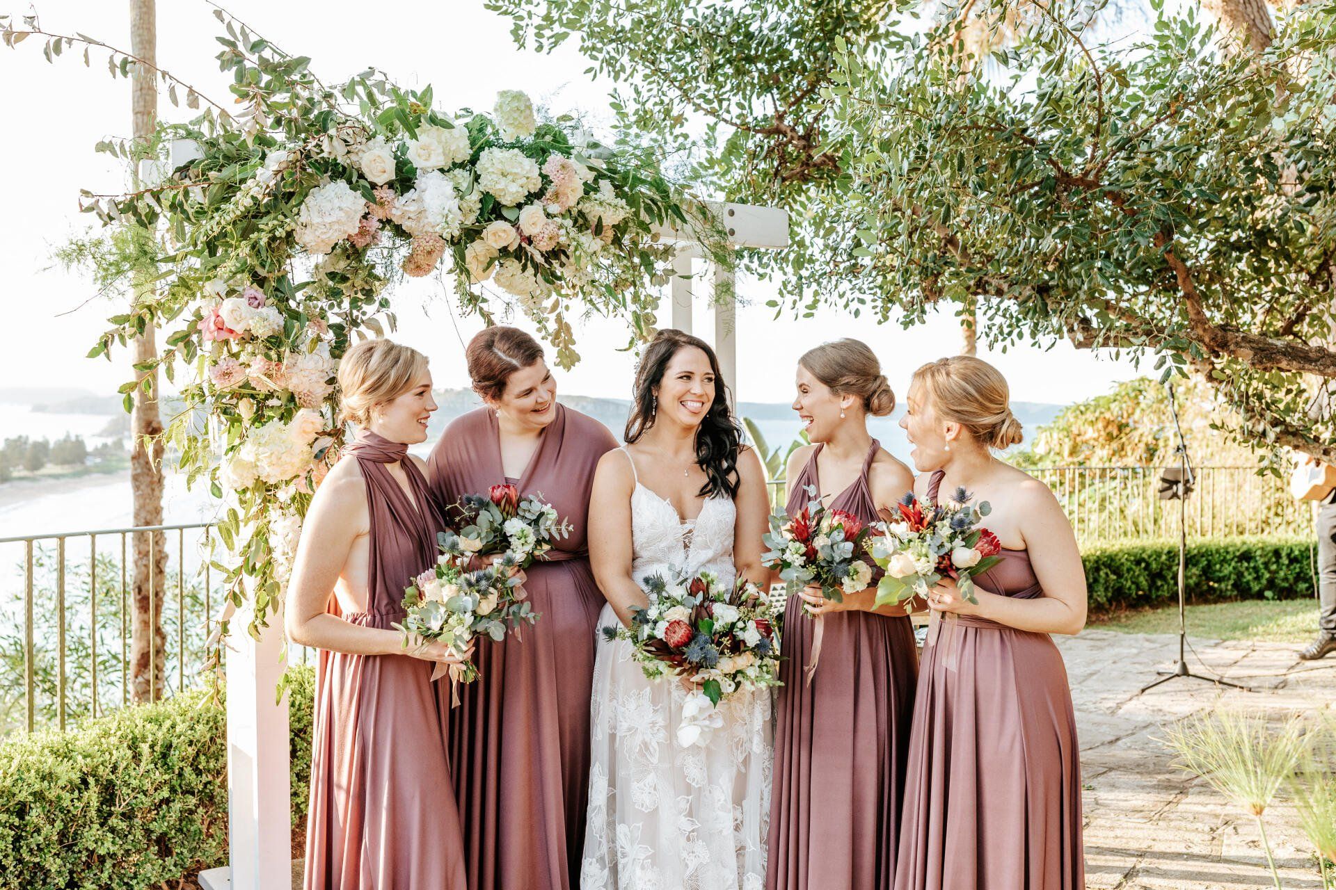 A bride and her bridesmaids are posing for a picture under a floral arch.