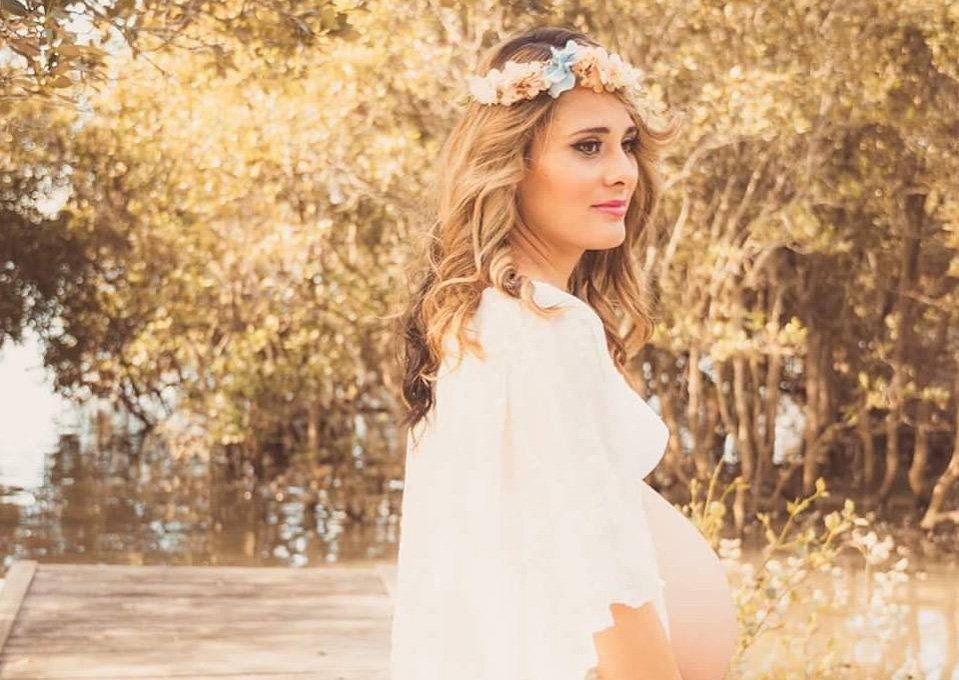 A pregnant woman wearing a flower crown is standing on a dock.