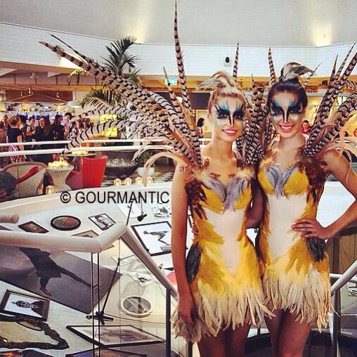 Two women in feathered costumes stand next to each other in front of a sign that says gourmantic