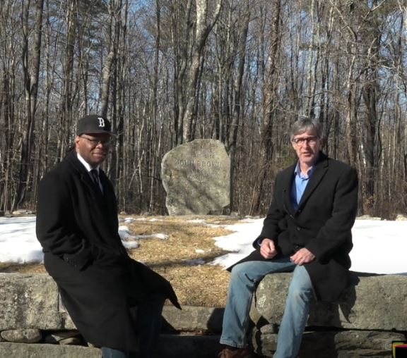 Two men sitting on a stone wall with one wearing a hat with the letter b on it