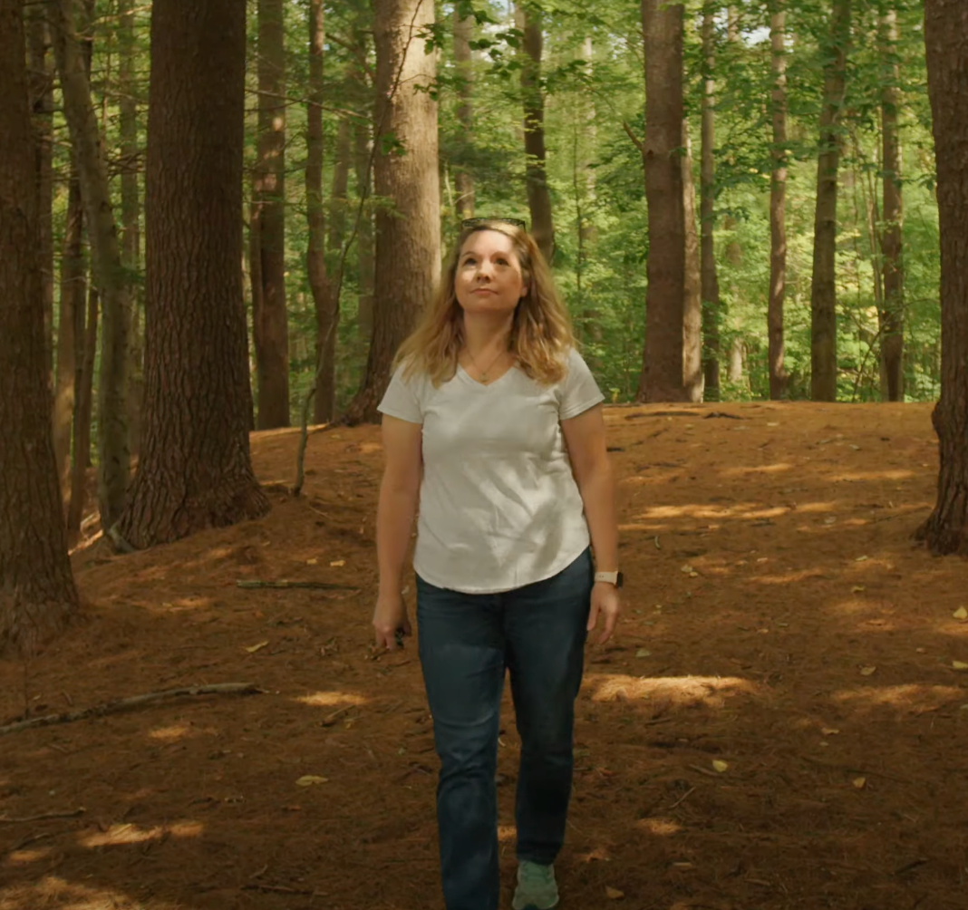 A woman in a white shirt and blue jeans is walking through a forest