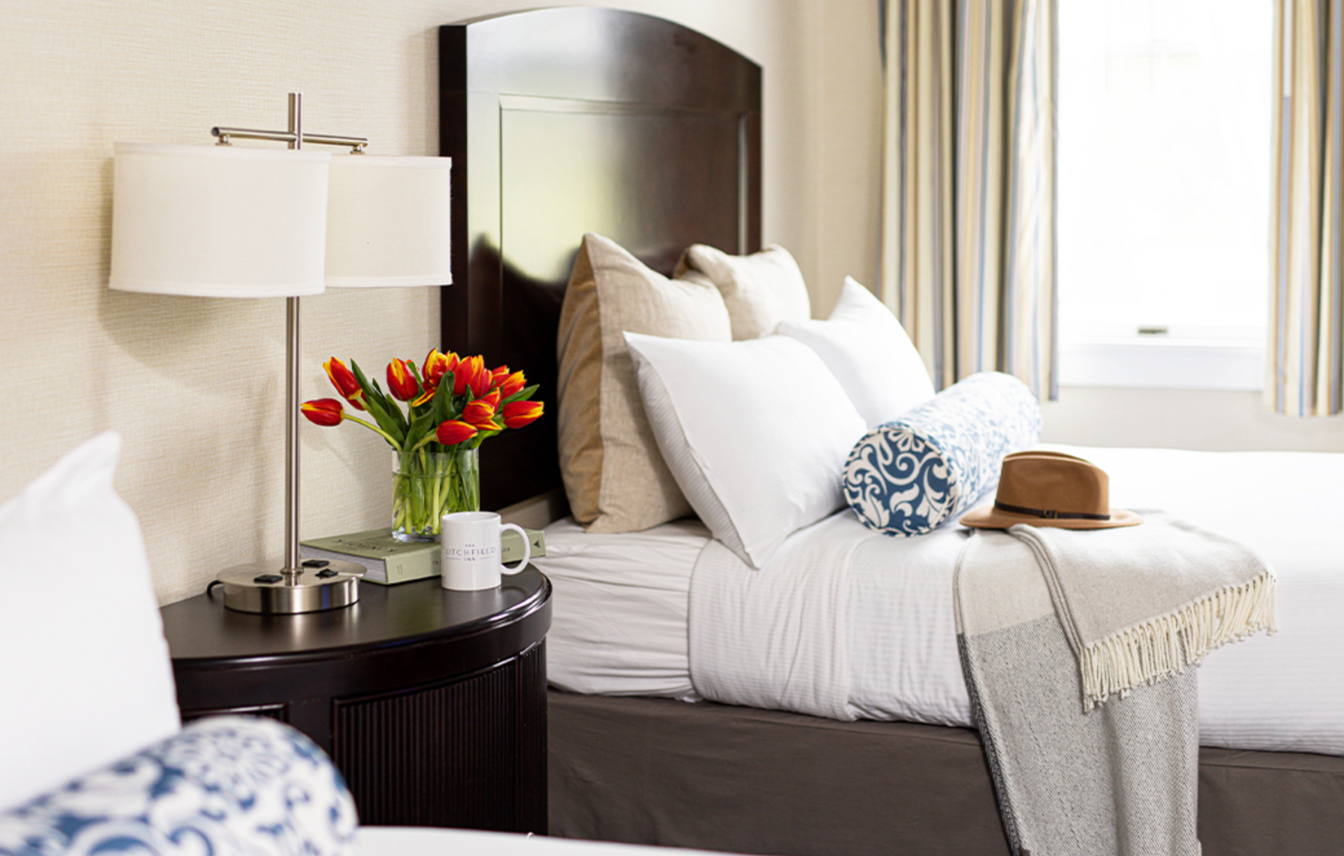 A hotel room with a bed , nightstand , lamp and a vase of flowers.