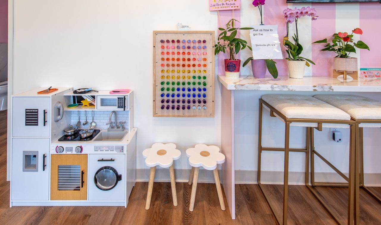 A play kitchen and two flower-shaped stools sit in front of a white wall, next to a counter with potted plants and seating.