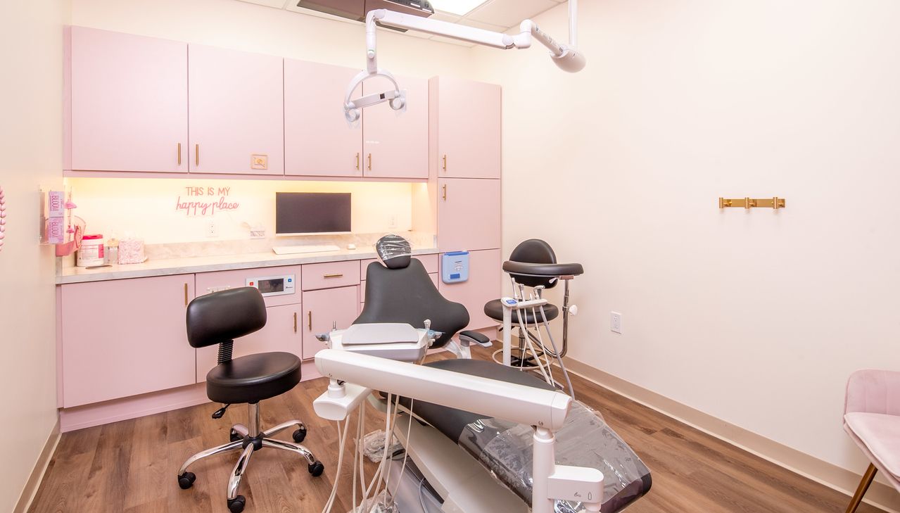 An exam room with a dental chair, medical equipment, and black stools against light pink cabinetry and wood flooring.